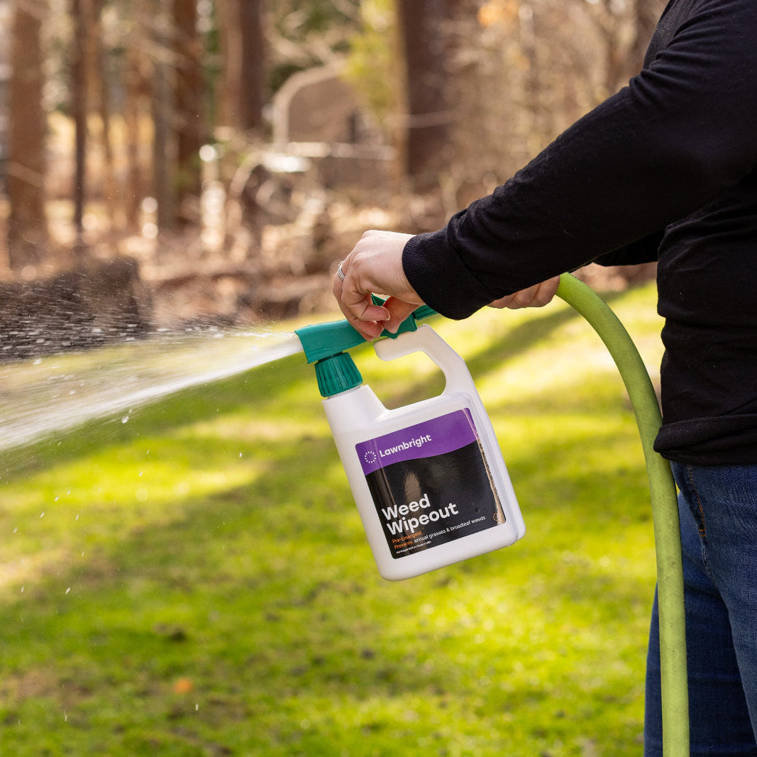 Person holding a bottle labeled 'Weed Out' over a garden hose with a grassy area and trees in the background.