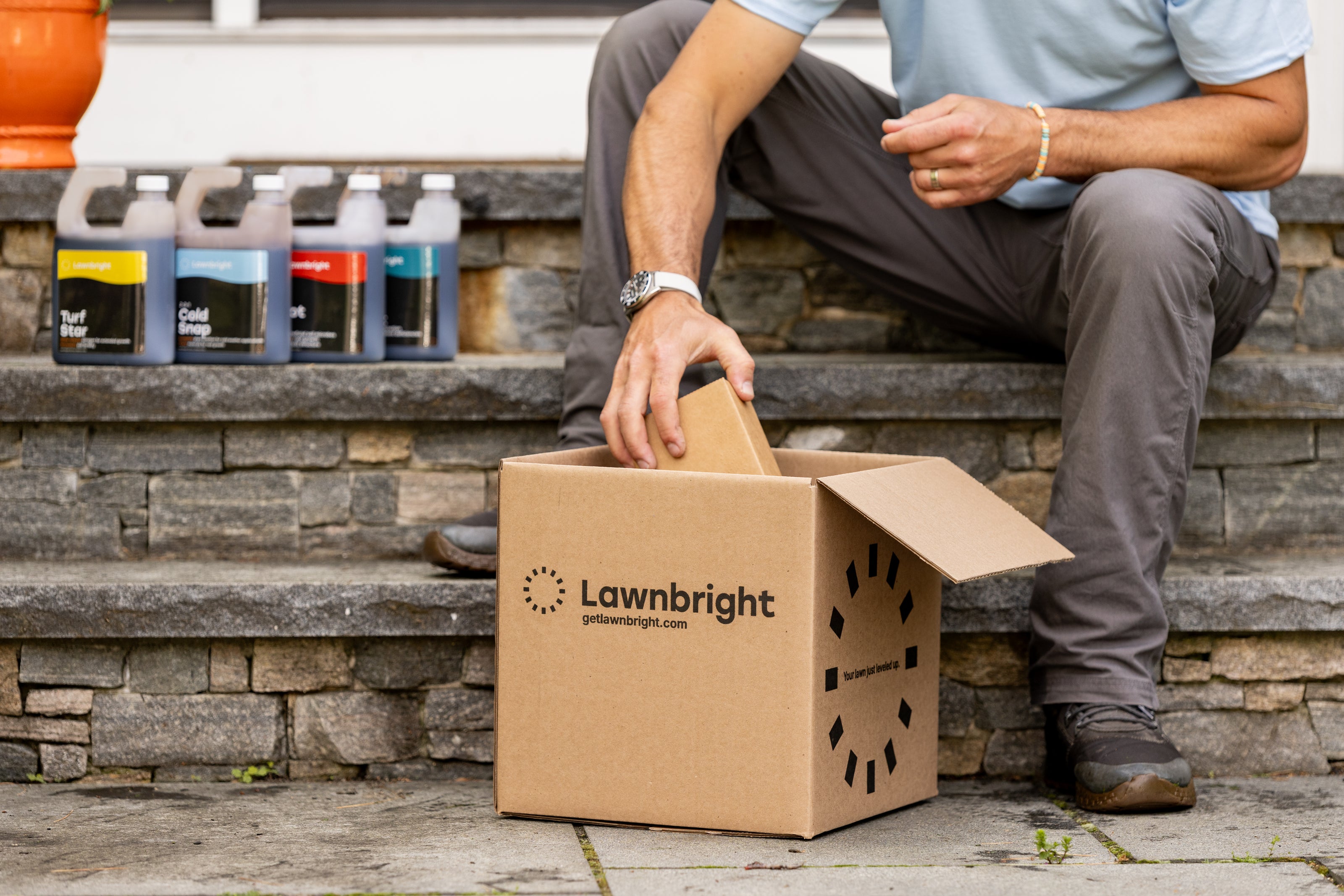 Person sitting on front steps opening a box labeled Lawnbright, with several bottles next to them.