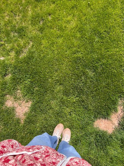 A top-down view of a person standing on green grass, wearing blue jeans, beige sneakers, and a red-and-white patterned top.