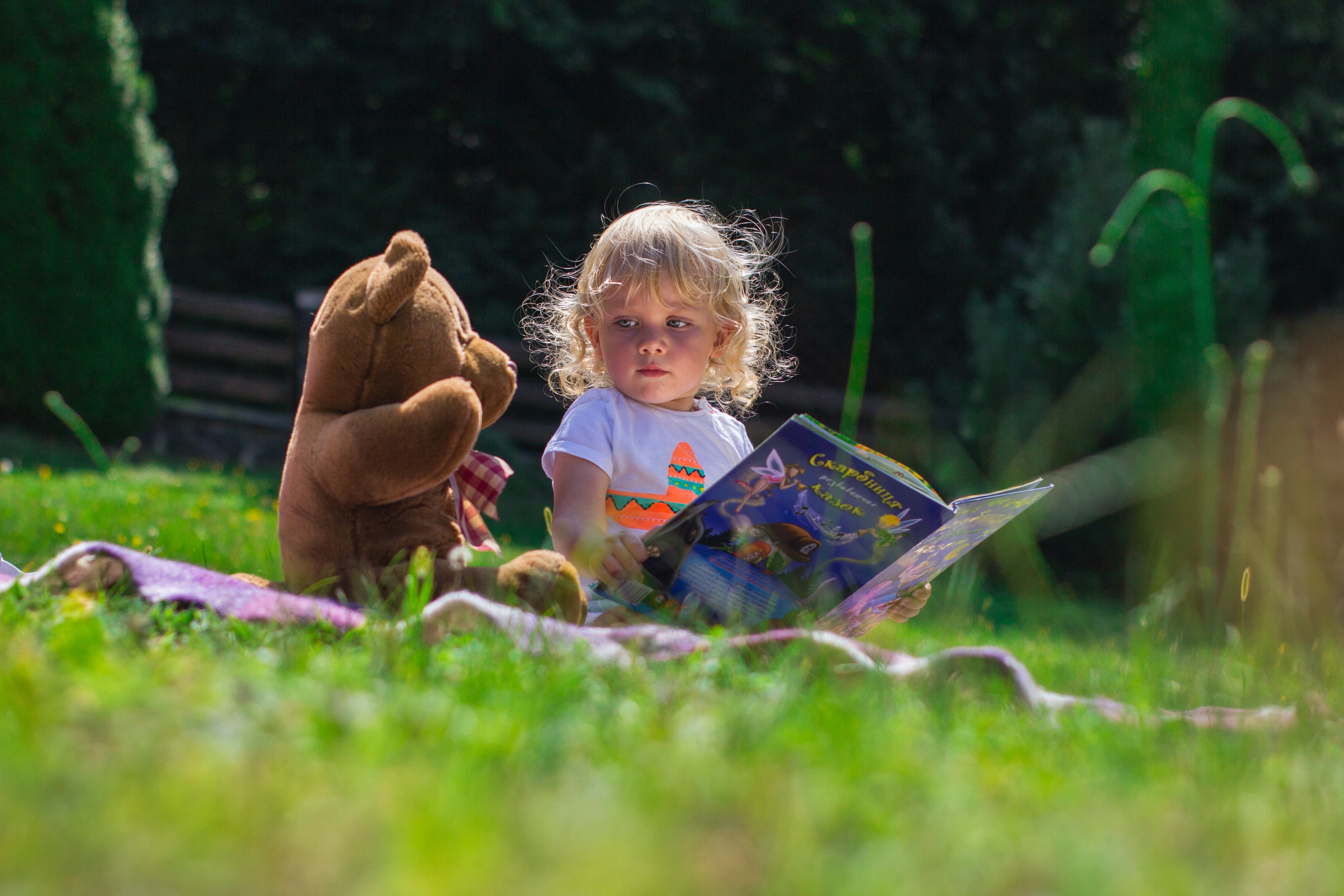 Small child on a lawn with a teddy bear, reading a book.